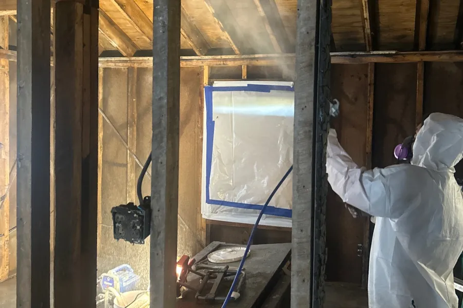 A person in a white hazmat suit and purple respirator sprays a mist on a charred wooden frame in a house under renovation.